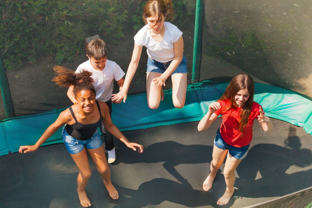 Top view of four friends, teenage girls and boys, bouncing on the outdoor trampoline in summer
